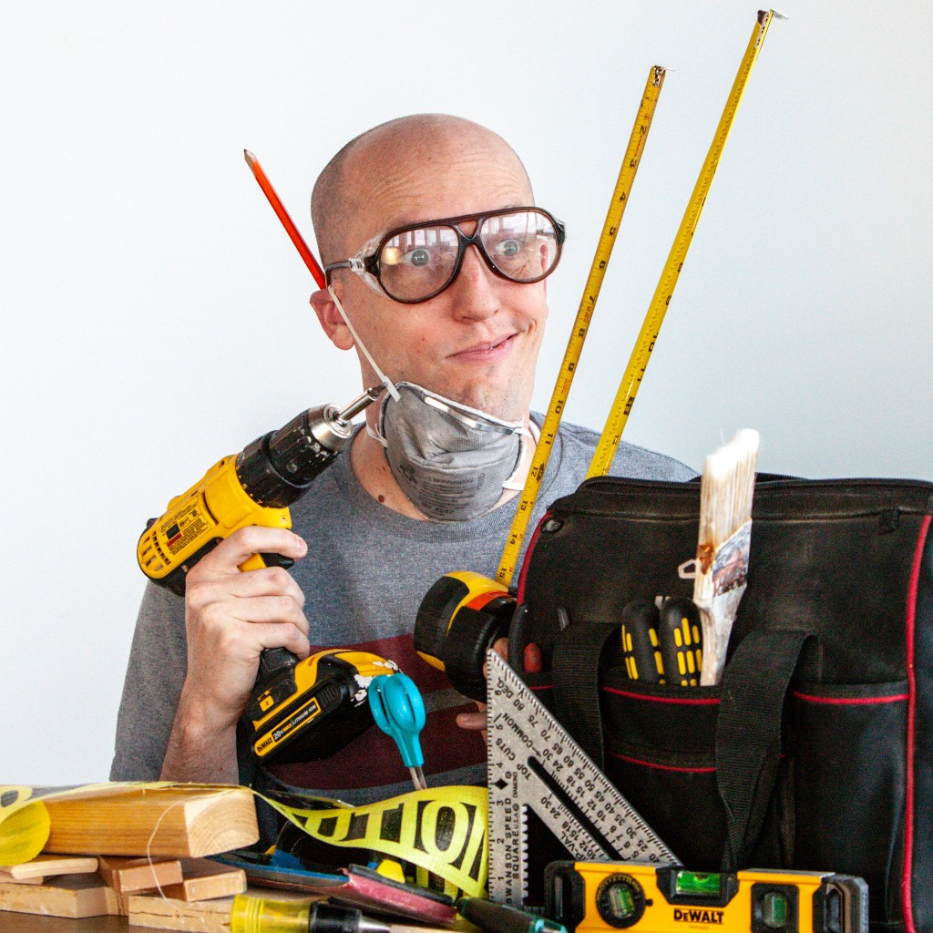 Founder of Family Quote Board in safety goggles with a dust mask around his neck, holding a drill amid DIY tools